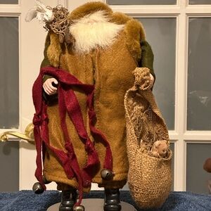 Shaggy haired and bearded Santa wears a brown cape and carries a burlap sack.